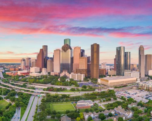 Aerial view of Houston skyline at sunset with vibrant skies, skyscrapers, highways, and green parks.