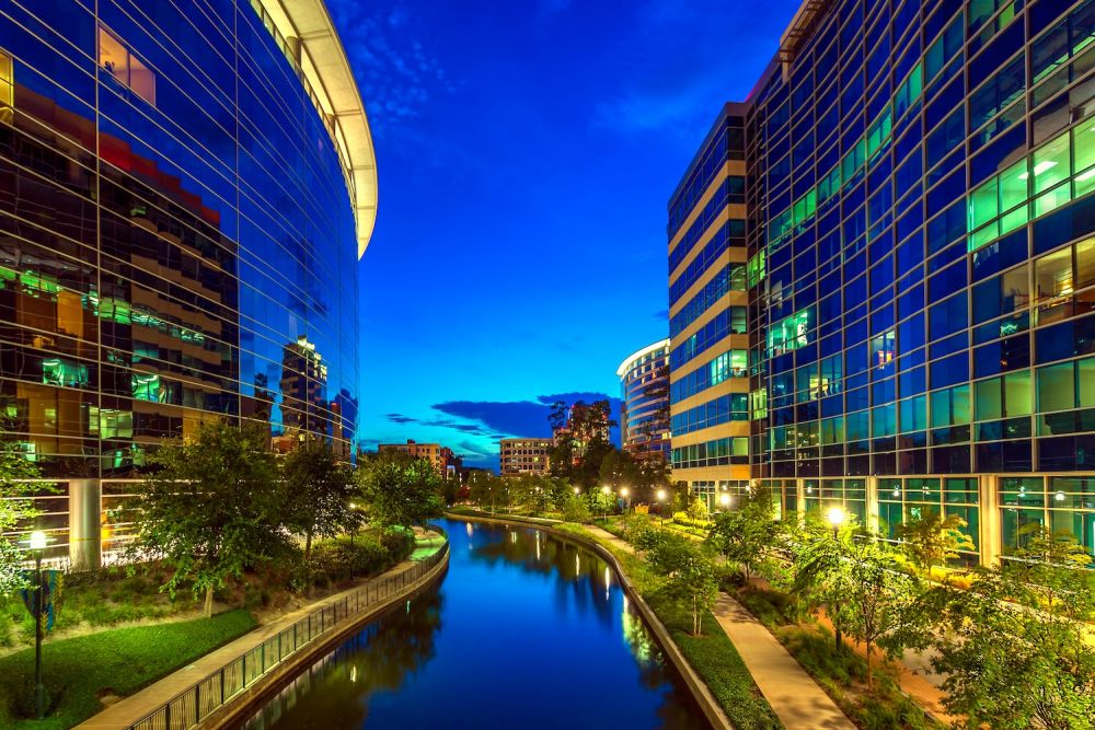 Night view of urban canal walkway flanked by modern glass buildings and vibrant city lights.