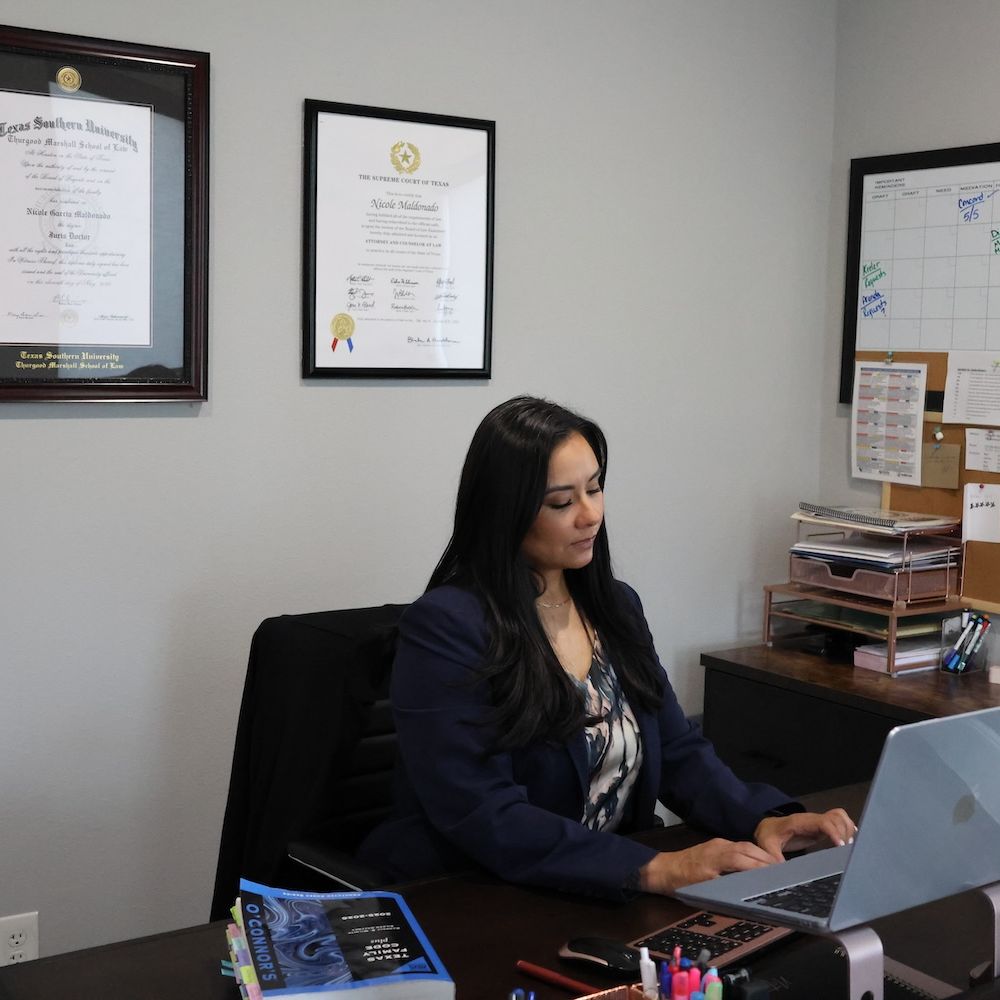 Nicole Maldonado working at desk with laptop, surrounded by diplomas and office supplies.