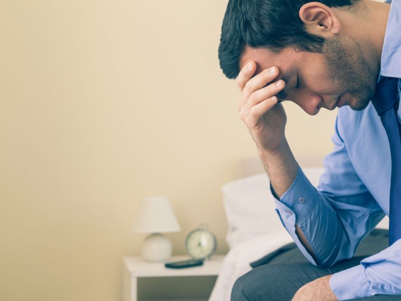 Man in blue shirt sitting on bed with head in hand, appearing stressed.