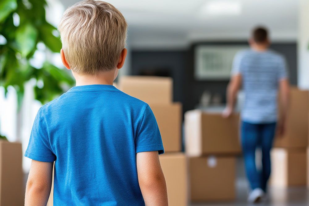 Child in blue shirt watches an adult moving boxes in a house.