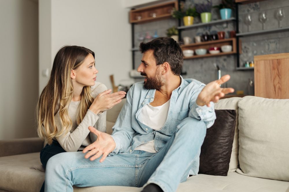 Couple arguing on a sofa in a living room, woman gesturing with her hands as she talks to the man.