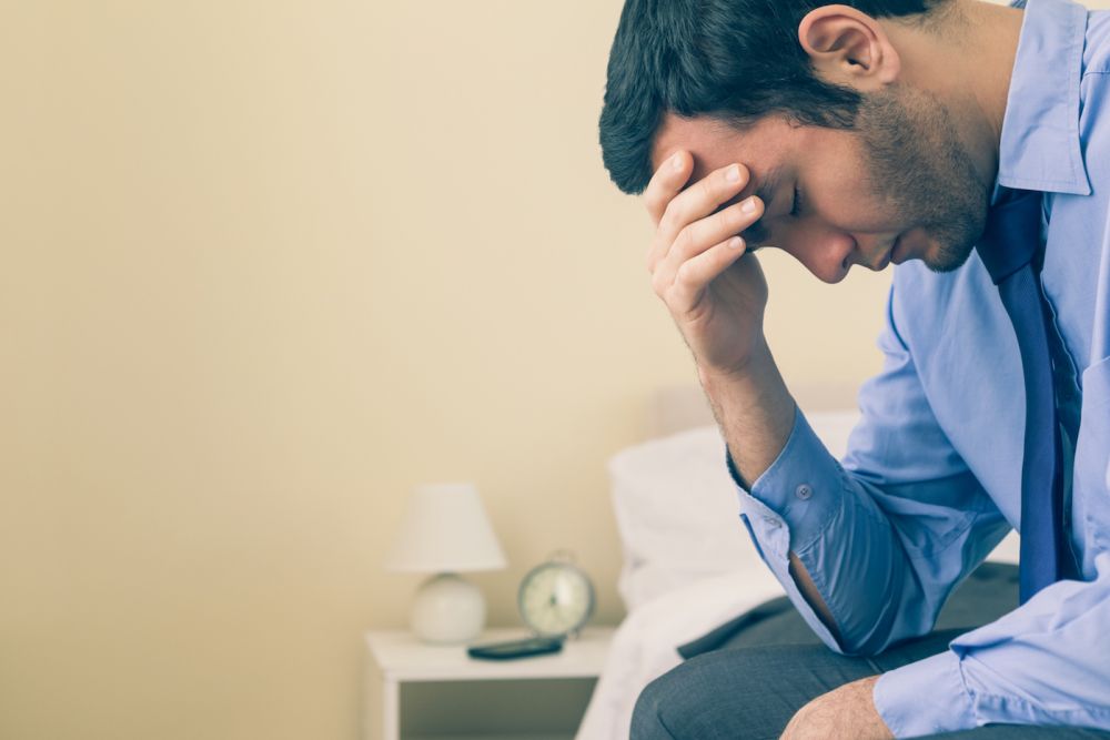 Man in blue shirt sitting on bed with head in hand, appearing stressed.