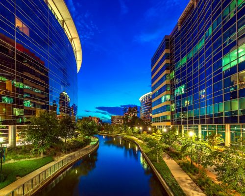 Night view of urban canal walkway flanked by modern glass buildings and vibrant city lights.
