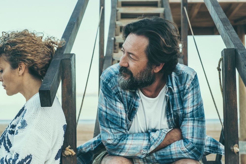 Couple sitting back to back on wooden structure, expressing tension and distance, with a blurred beach background.