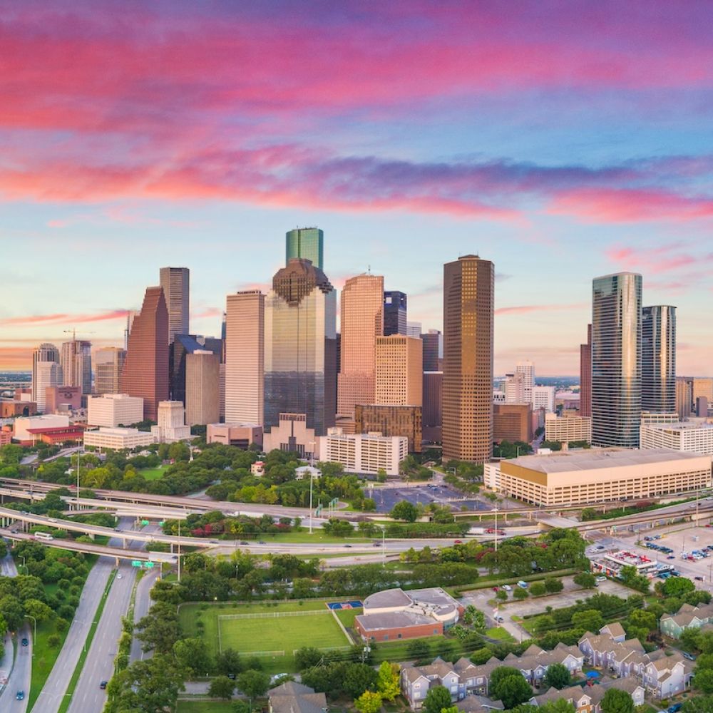 Aerial view of Houston skyline at sunset with vibrant skies, skyscrapers, highways, and green parks.