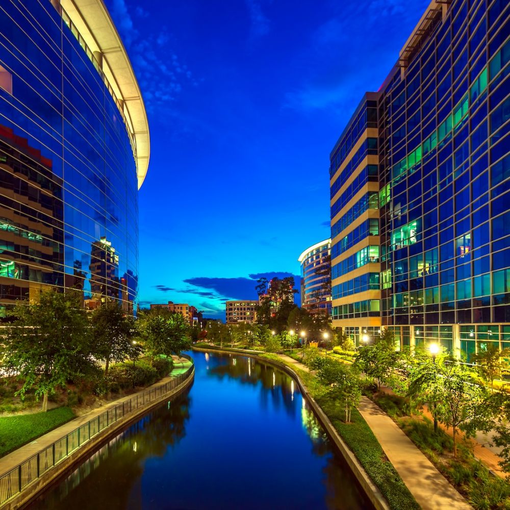 Night view of urban canal walkway flanked by modern glass buildings and vibrant city lights.