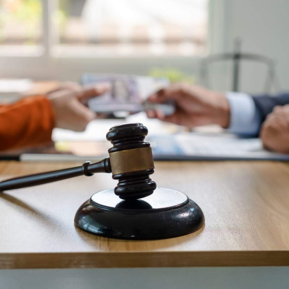 Close-up of a wooden gavel on a desk with blurred hands of two people shaking hands in the background, suggesting a legal agreement or decision.