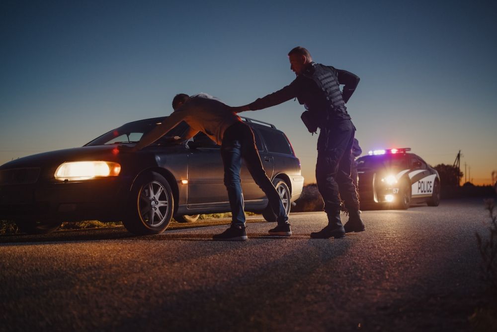 Police officer searches car driver during evening traffic stop, with patrol car flashing lights in background.