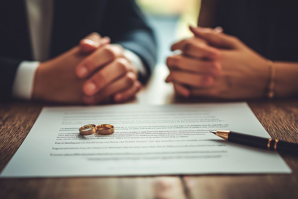 Two people sit facing each other with wedding rings and a divorce agreement on the table.