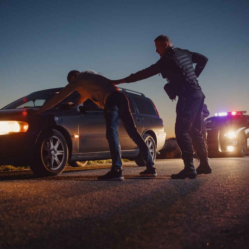 Police officer searches car driver during evening traffic stop, with patrol car flashing lights in background.