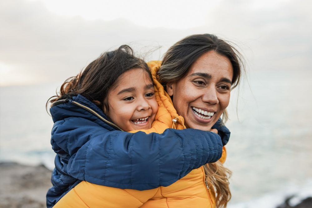 Mother and child smiling by the sea, bundled in colorful jackets, enjoying a joyful outdoor moment.