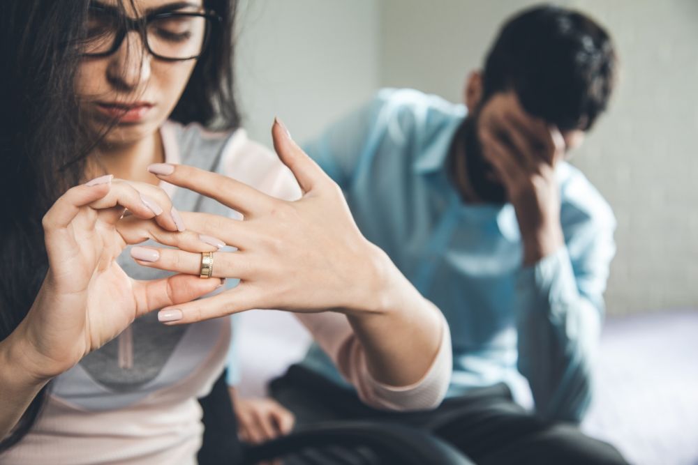 Woman removing wedding ring, man in background, emotional couple conflict concept.
