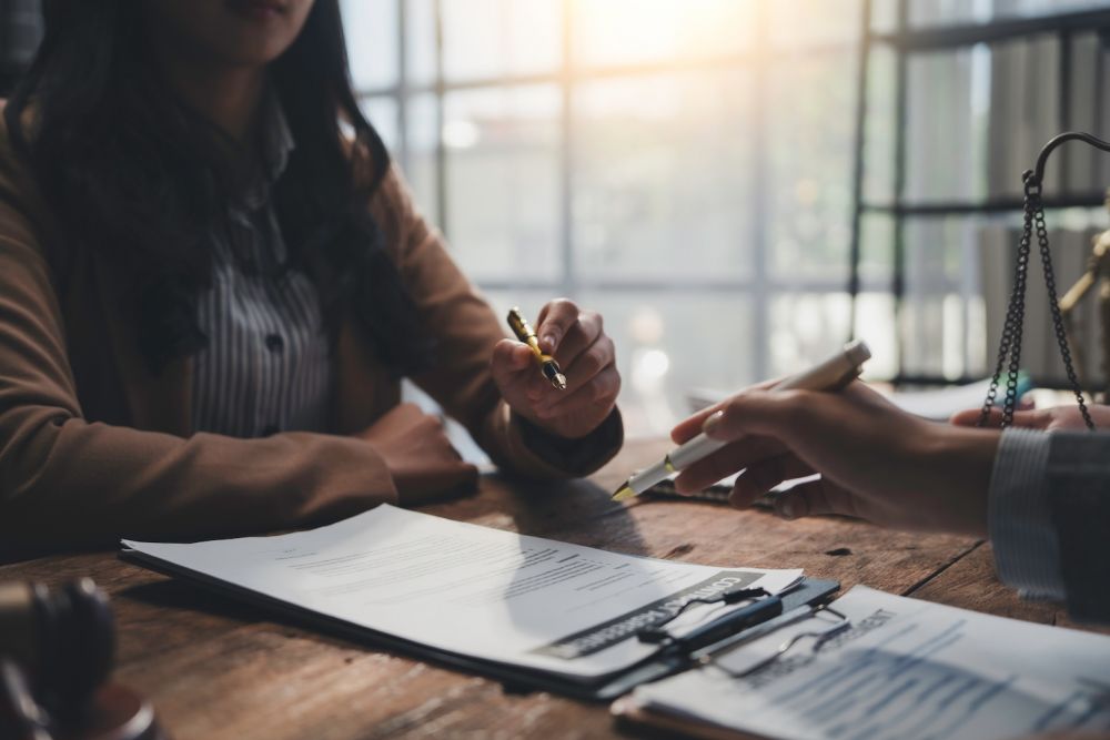 Two people discuss and review documents at a wooden table in an office, with pens and paper in hand.