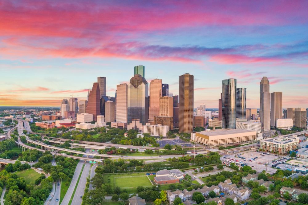 Aerial view of Houston skyline at sunset with vibrant skies, skyscrapers, highways, and green parks.