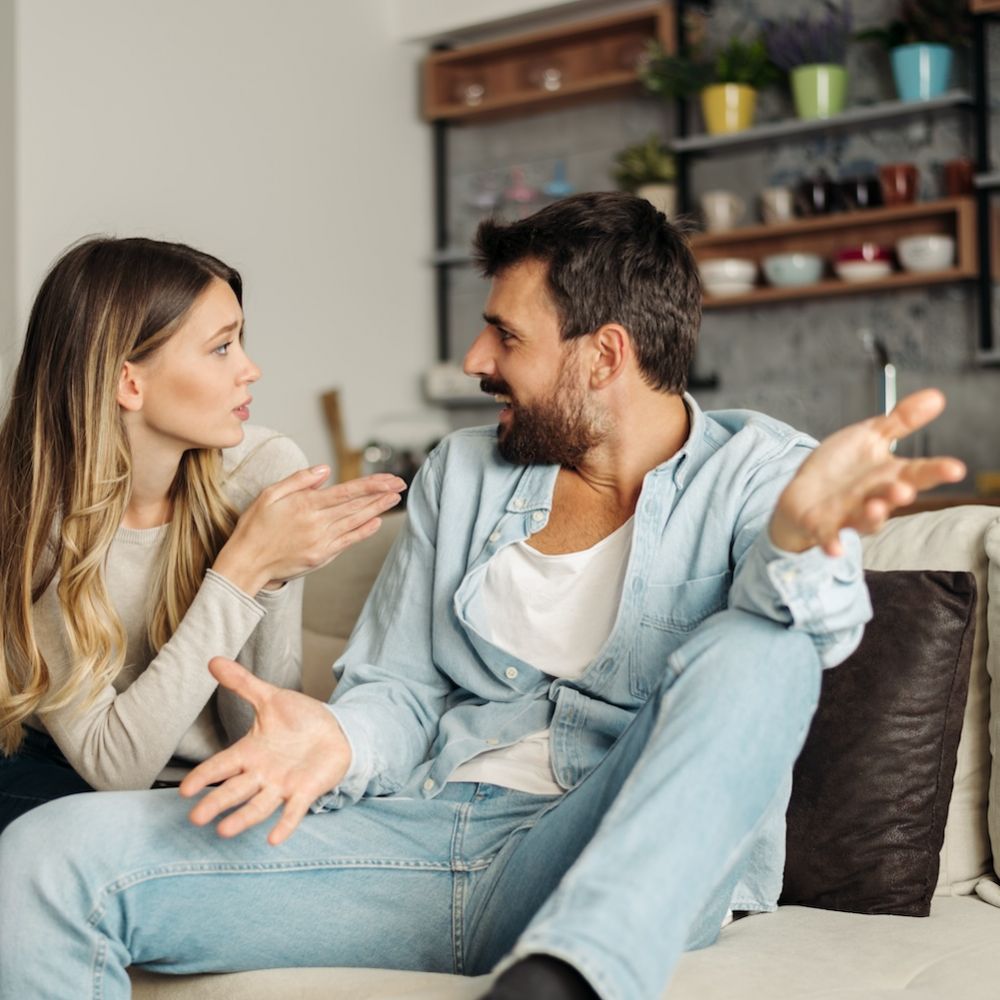 Couple arguing on a sofa in a living room, woman gesturing with her hands as she talks to the man.