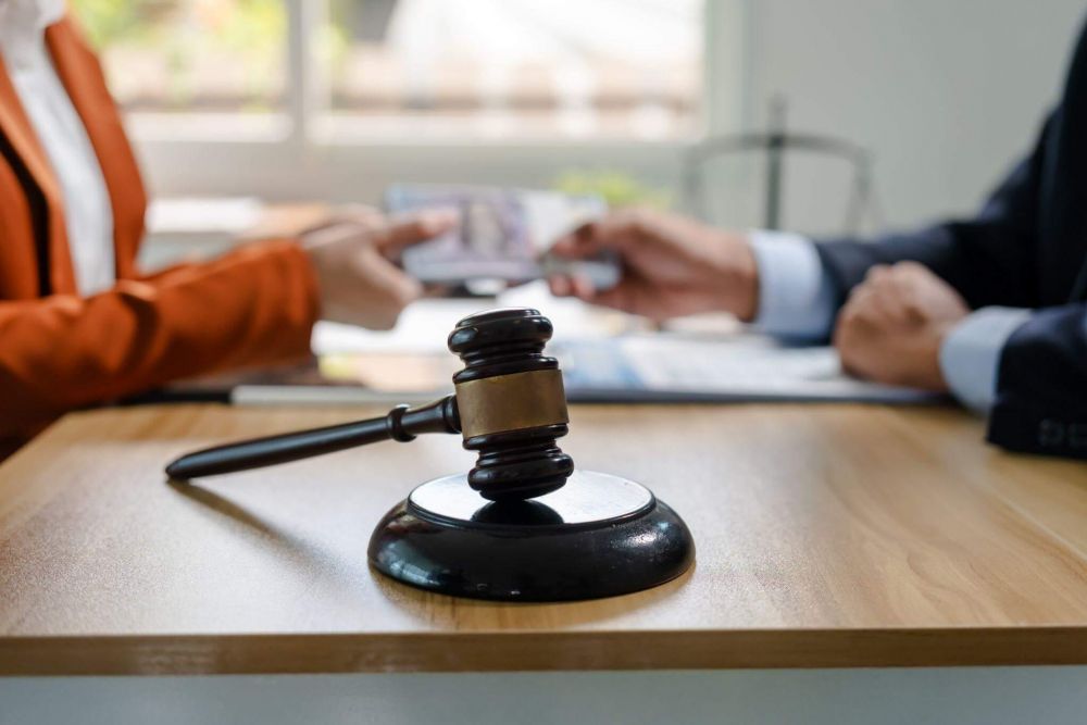 Close-up of a wooden gavel on a desk with blurred hands of two people shaking hands in the background, suggesting a legal agreement or decision.