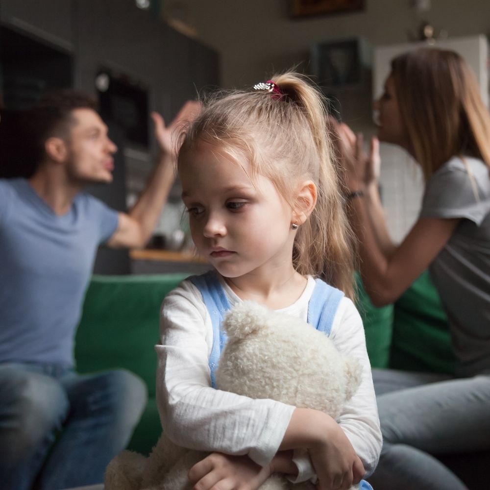 Young girl holding teddy bear, looks upset as parents argue in background.