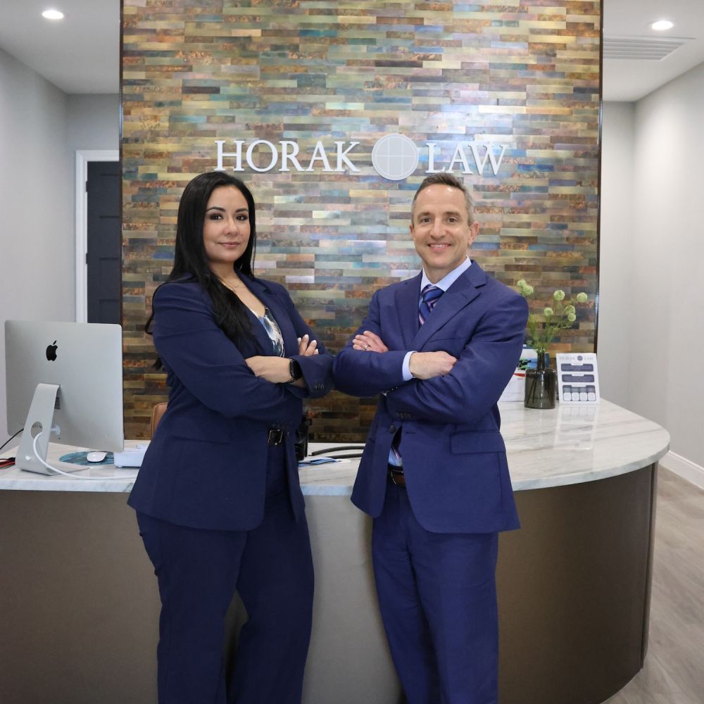 Matthew P. Horak and Nicole Maldonado in blue suits pose with arms crossed at a law firm reception, in front of a colorful stone wall with a Horak Law logo.