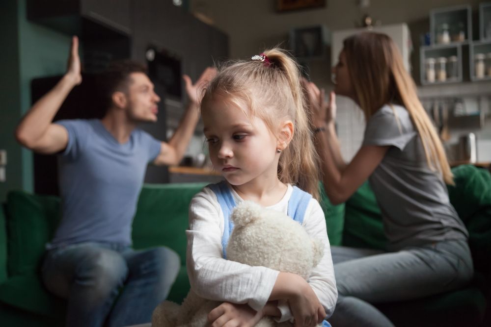 Young girl holding teddy bear, looks upset as parents argue in background.